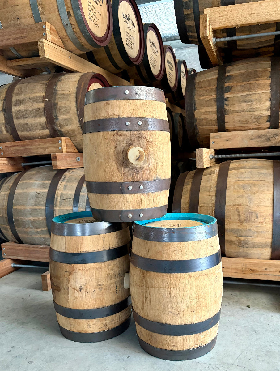 Several wooden wine barrels stacked on wooden pallets in a warehouse setting. The barrels have natural light brown wood staves, dark metal hoops, and one barrel features a turquoise blue interior visible at the top.