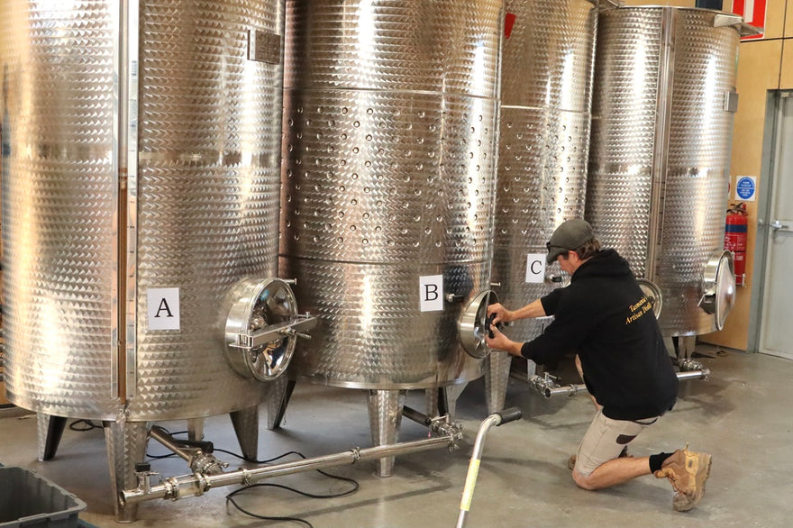 A man in a black hoodie and khaki pants kneels beside large stainless steel fermentation tanks labeled A, B, and C in a brewery setting.