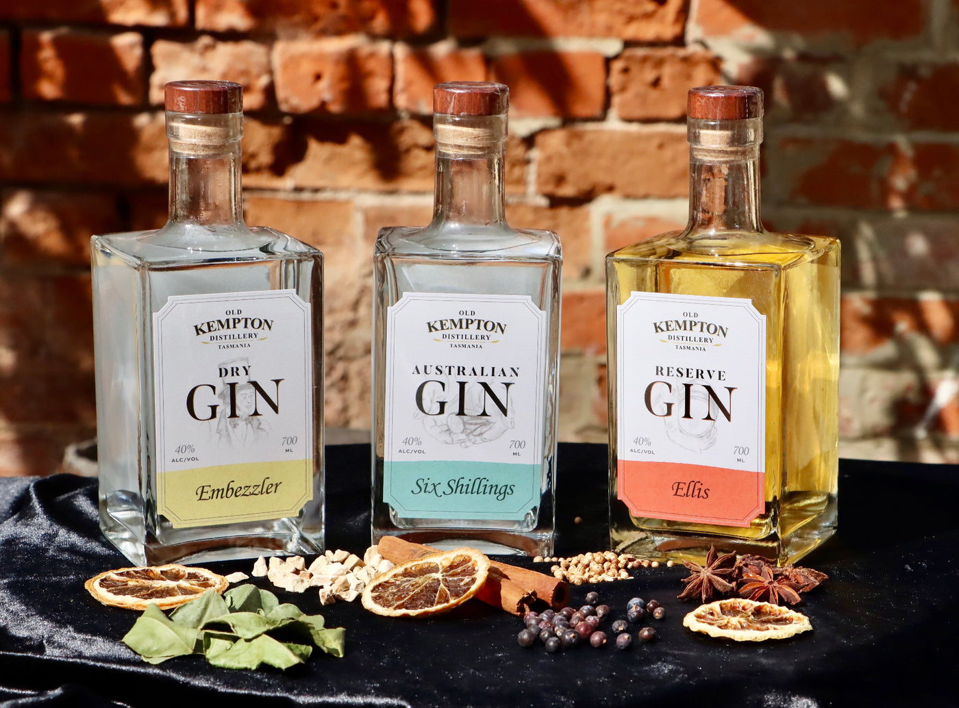 Three bottles of Kempton Distillery gin with decorative elements on a dark surface against a brick wall.