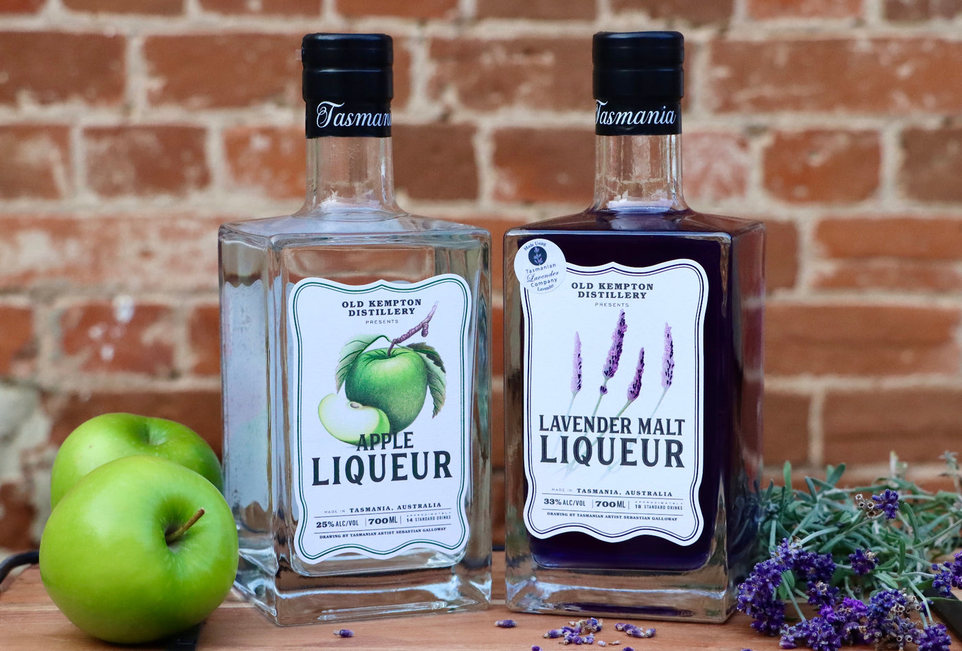Two bottles of Tasmanian Old Kempton Distillery liqueurs with apples and lavender on a wooden surface against a brick wall.