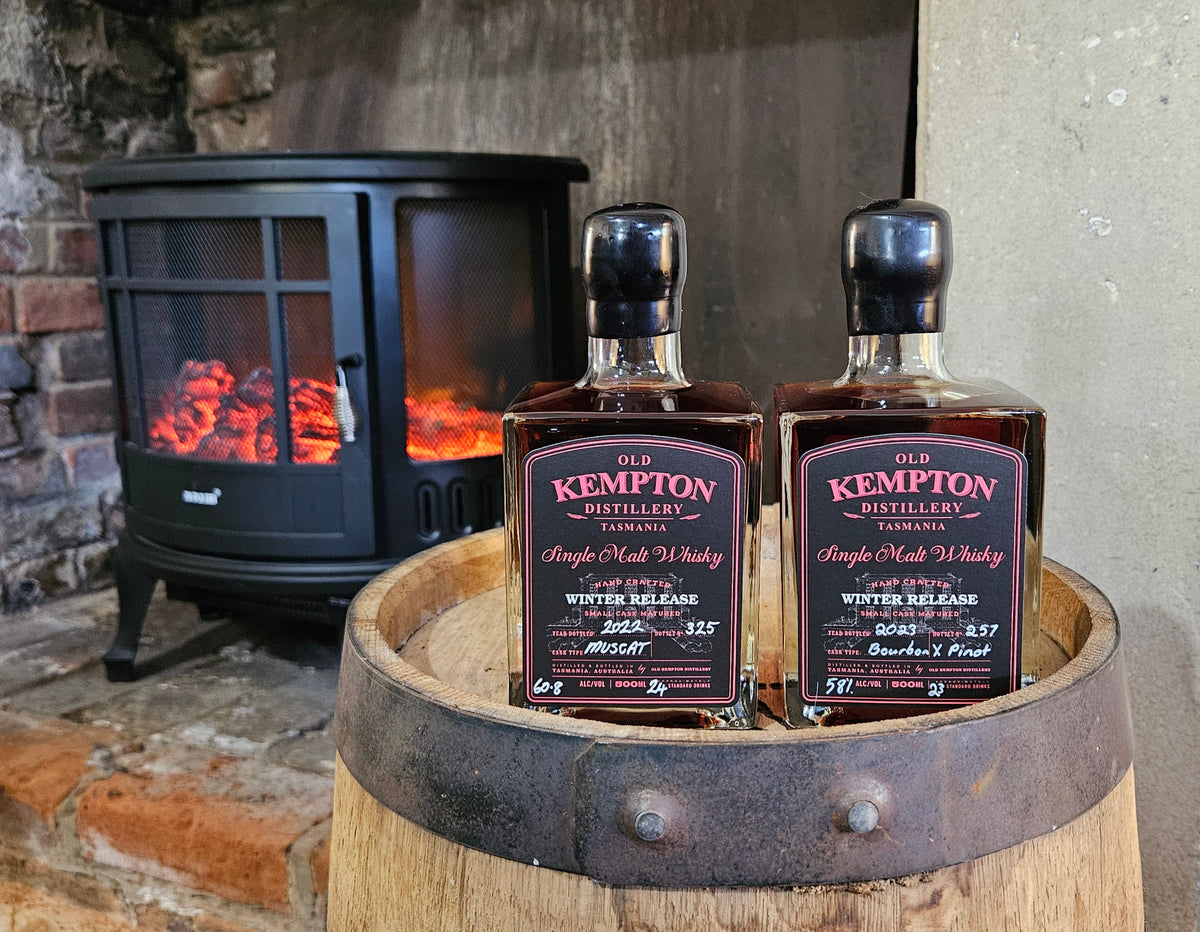 Two bottles of Kempton Distilled Single Malt Whisky placed on a wooden barrel in front of a lit fireplace.