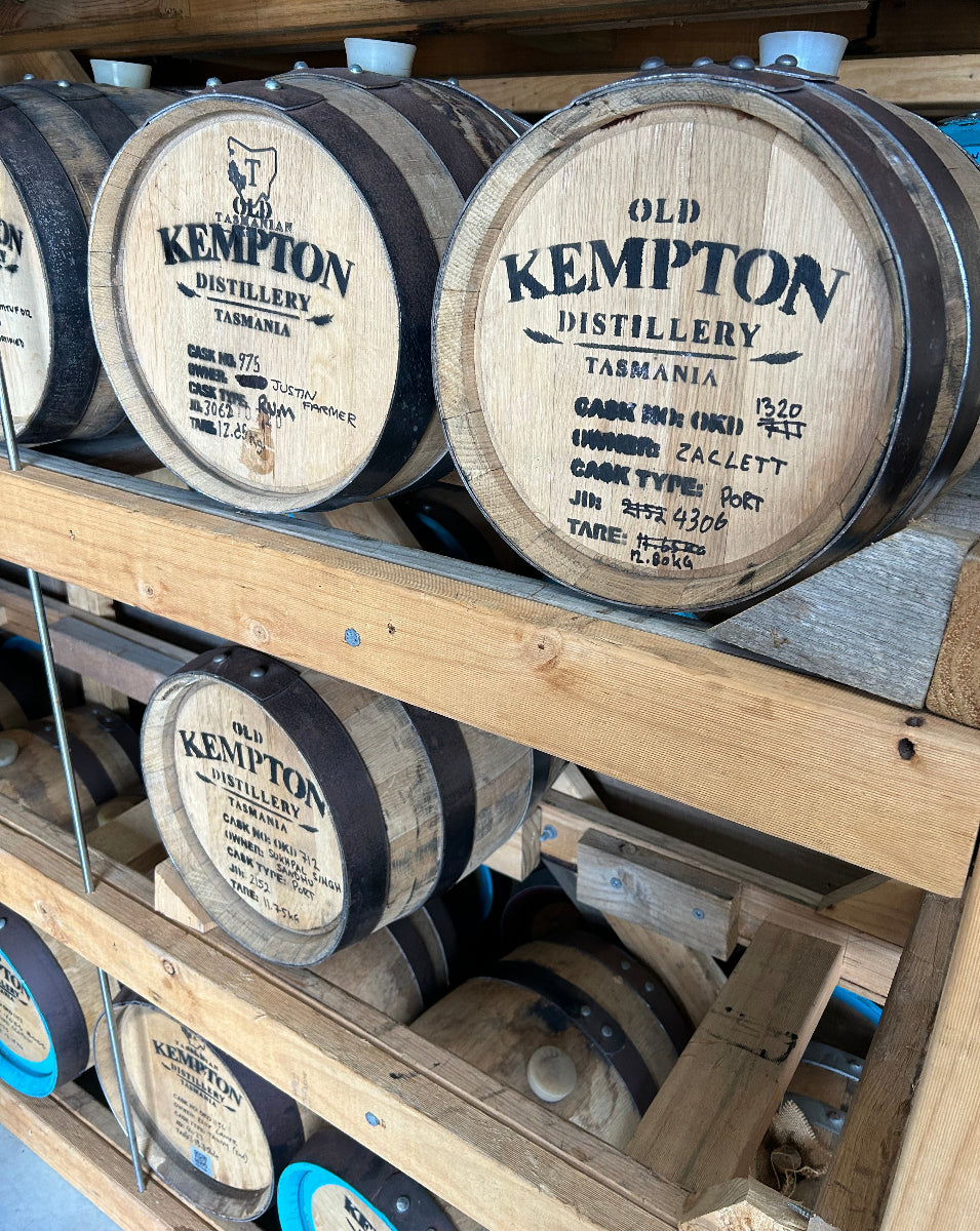 Wooden barrels from Old Kempton Distillery in Tasmania, stacked on wooden shelves. The barrels feature black metal hoops and printed black labels with the company name and barrel details.